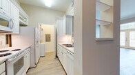 This single-floor residence offers a thoughtfully designed layout with a clean, neutral palette and a variety of functional spaces. The galley-style kitchen is outfitted with white cabinetry extending to the ceiling, a double stainless steel sink, white appliances including a refrigerator with ice dispenser, electric range, and built-in microwave, all set atop light hardwood-style flooring. A dedicated laundry area is conveniently accessible directly from the kitchen. The living area features light hardwood-style flooring, crown molding, and a pass-through opening to the kitchen, complemented by built-in shelving along one wall. The entry area is highlighted by elegant French doors flanked by sidelights and a transom window, flooding the space with natural light. The carpeted bedrooms are generously sized; one features a ceiling fan, large windows with natural views, and ample wall space, while another boasts elevated ceilings, a transom window above the doorway, and access to a spacious walk-in closet. The bathroom presents a long white vanity with an extended countertop, a large mirror, pendant lighting, tile flooring, and a full soaking tub with shower. Throughout the residence, warm beige tones and light finishes create a bright, welcoming ambiance.
