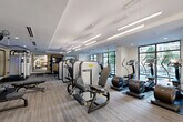Located at 2303 Mid Lane in Houston's prestigious River Oaks corridor, this modern fitness center offers a thoughtfully designed, single-floor layout that accommodates a full range of workout needs. The space is bathed in natural light streaming through large floor-to-ceiling windows that frame views of the surrounding greenery, creating an open and energizing atmosphere.

The fitness floor is finished with durable, textured carpet tiles and features a tray ceiling with sleek linear pendant lighting, lending the space a polished, contemporary aesthetic. The equipment selection is both varied and high-end, including multiple treadmills, elliptical trainers, stair climbers, and stationary bikes for cardiovascular conditioning. A dedicated strength training zone houses a multi-functional cable machine, weight bench, and a well-stocked dumbbell rack arranged alongside a large framed mirror and a wall-mounted flat-screen television.

TRX suspension straps are anchored near the windows, offering functional training options. Strategically placed mirrors throughout the space enhance the sense of depth and allow users to monitor their form. Near the entrance, a built-in shelving unit with towel service adds a convenient, amenity-focused touch. The fitness center is accessible via both a glass-paneled interior door and a direct exterior entry, ensuring easy access for users.