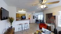 This single-floor residence offers an open-concept layout that seamlessly connects the kitchen, dining, and living areas. The kitchen is equipped with warm wood-toned cabinetry, a tile backsplash, and a peninsula counter with bar seating for two, making it a functional and social hub. A fluorescent light fixture illuminates the kitchen workspace, while flush-mount ceiling lights provide additional brightness throughout the common areas.

The living space flows naturally from the kitchen, featuring light wood-style laminate flooring that runs throughout and ties the areas together visually. A ceiling fan with an integrated light kit is centrally positioned, promoting air circulation and adding to the room's comfort. The living area accommodates seating and a coffee table, while a window dressed with white shutters allows natural light to filter into the dining zone, where a table and chairs are neatly arranged.

A textured ceiling is present throughout the space. Doorways visible from the main area suggest access to additional rooms. The overall layout is practical and well-connected, offering a cohesive living environment suited for everyday use.