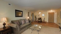 This single-floor residence offers a thoughtfully designed layout that emphasizes open, connected living. The living area is warm and inviting, featuring plush carpeting underfoot and neutral-toned walls that create a calm, welcoming ambiance. A decorative framed mirror adds depth to the space, while a classic table lamp provides soft, ambient lighting. The living area flows seamlessly into the dining space, which is defined by rich hardwood-style flooring and anchored by a traditional dining set arranged atop a patterned area rug. A ceiling fan with an integrated light fixture serves both the dining and adjacent areas, ensuring comfort and functionality throughout. The kitchen is accessible directly from the dining area, with a stainless steel refrigerator visible through the connecting passage, hinting at a practical and well-equipped culinary space. Decorative touches such as potted plants and framed wall art lend a lived-in, homey character to the interior. The open sightlines between rooms enhance the sense of spaciousness, making the layout feel airy and easy to navigate. Overall, the space presents a cohesive, comfortable environment with a classic residential aesthetic and attention to detail in both design and furnishings.
