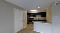 This single-story space centers around a well-appointed kitchen that serves as its standout feature. The kitchen is outfitted with rich, dark cabinetry that extends to the upper and lower sections of the walls, providing generous storage throughout. Granite countertops with subtle veining add a polished, durable surface for meal preparation and everyday use. Stainless steel appliances, including a gas range, microwave, refrigerator, and sink, lend a contemporary, cohesive aesthetic to the space.

A peninsula with a light-colored countertop extends from the kitchen, creating a natural divide between the kitchen and the adjacent carpeted living area. This transitional element offers additional counter space and serves as a casual dining or gathering spot. The entryway, positioned near the kitchen, features a classic six-panel white door that contrasts cleanly with the neutral beige walls.

Recessed lighting and a flush-mount ceiling fixture provide ample illumination throughout the kitchen area. Light wood-look flooring anchors the kitchen, while carpeting transitions into the living space, adding warmth. The overall ambiance is modern and functional, with a neutral palette that complements the bold cabinetry.