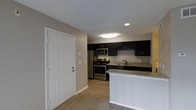 This single-story space centers around a well-appointed kitchen that serves as its standout feature. The kitchen is outfitted with rich, dark cabinetry that extends to the upper and lower sections of the walls, providing generous storage throughout. Granite countertops with subtle veining add a polished, durable surface for meal preparation and everyday use. Stainless steel appliances, including a gas range, microwave, refrigerator, and sink, lend a contemporary, cohesive aesthetic to the space.

A peninsula with a light-colored countertop extends from the kitchen, creating a natural divide between the kitchen and the adjacent carpeted living area. This transitional element offers additional counter space and serves as a casual dining or gathering spot. The entryway, positioned near the kitchen, features a classic six-panel white door that contrasts cleanly with the neutral beige walls.

Recessed lighting and a flush-mount ceiling fixture provide ample illumination throughout the kitchen area. Light wood-look flooring anchors the kitchen, while carpeting transitions into the living space, adding warmth. The overall ambiance is modern and functional, with a neutral palette that complements the bold cabinetry.