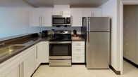 This thoughtfully designed two-bedroom, one-bathroom residence spans a single floor and offers a functional, comfortable layout ideal for everyday living. The kitchen features white shaker-style cabinetry, dark granite-style countertops, and a full suite of stainless steel appliances, including a range, over-the-range microwave, and refrigerator, all set atop light tile flooring. The living area provides a relaxed setting with carpeted floors and ample space for seating. Both bedrooms are carpeted and similarly appointed, each accommodating two twin beds with dark upholstered headboards, a shared nightstand with a lamp, and windows that invite natural light and views of the surrounding outdoor scenery. One bedroom also features a dedicated closet for added storage. The bathroom is well-appointed with white cabinetry, a large vanity mirror, white countertops, and a shower-tub combination, with clean light tile flooring throughout. A separate vanity area with additional cabinet storage provides extra convenience. The unit's neutral color palette and consistent finishes throughout create a cohesive and welcoming atmosphere from room to room.