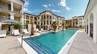 Located at 12701 Eastlake Blvd in El Paso, TX, this outdoor pool amenity area showcases a beautifully designed resort-style retreat. The centerpiece is a generously sized, crystal-clear swimming pool with vivid turquoise water, complemented by a separate spa featuring a waterfall feature. The pool deck is finished in light-colored concrete and lined with white lounge chairs, providing ample space for sunbathing beneath the West Texas sky. Tall, mature palm trees are strategically placed throughout the deck, lending a tropical ambiance to the space. The area is enclosed by a decorative wrought-iron fence, ensuring security while maintaining an open, airy feel. Surrounding the pool on multiple sides are multi-story residential buildings designed in a Spanish Colonial Revival style, characterized by white stucco exteriors, terracotta tile rooflines, columned arcades, and private balconies. A single-story clubhouse with elegant arched colonnades and large glass windows faces the pool directly, offering a seamless indoor-outdoor connection. An American flag is prominently displayed atop the clubhouse roofline. The overall ambiance is upscale and resort-like, ideal for relaxation and outdoor recreation in El Paso's sunny climate.