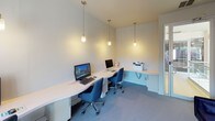 This single-floor business center is thoughtfully designed to support productivity in a comfortable, professional environment. The space is anchored by a generously sized, built-in wrap-around desk with a light wood finish that accommodates multiple workstations, each equipped with desktop computers and keyboards. Ergonomic navy blue office chairs complement the desk setup and tie into the room's cohesive color palette.

A series of sleek pendant lights hang from the tall ceilings, casting warm, focused illumination across the workspace. The soft blue-grey carpeting underfoot adds a sense of calm and sound absorption throughout the room. A printer station is conveniently integrated into the corner of the desk, making the space fully functional for a range of office needs.

Natural light filters into the room through large windows dressed with sheer curtains, offering views of the surrounding exterior while maintaining a bright and open atmosphere. A curved bay window adds architectural character to one end of the room.

At the far end of the space, a plush navy blue daybed sofa provides a comfortable lounge area beneath a striking large-scale abstract artwork in ocean-inspired blues and whites. This seating nook offers a relaxed counterpart to the more structured workstations, making the overall space versatile and inviting.