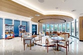 Located at The Breton in Bayonne, NJ, this elegant resident lounge offers a sophisticated and welcoming atmosphere designed for both relaxation and social interaction. The space is anchored by a striking circular lounge area beneath a warm wood-paneled curved ceiling detail, creating an intimate focal point within the open floor plan. Floor-to-ceiling windows wrap around the seating area, flooding the interior with natural light and offering views of the surrounding outdoors and waterfront.

The furnishings are carefully curated, featuring a curved cognac leather sofa, neutral upholstered chairs, and warm wood-toned round tables, all complementing the refined aesthetic. Polished marble-style flooring extends throughout, enhancing the bright and airy feel of the space. A built-in bar counter with high stools provides a casual spot for residents, while a coffee station near the entry adds everyday convenience.

The entrance is defined by natural wood cladding and a branded entry mat, establishing a polished first impression. A game area with arcade machines offers a playful counterpoint to the lounge's sophisticated design. Bold blue accent walls and contemporary artwork add color and visual interest throughout. Double glass doors open directly to an outdoor terrace, seamlessly connecting the interior to waterfront views and outdoor seating.