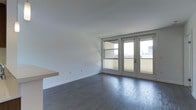 This contemporary single-floor space is defined by its clean, minimalist design and thoughtful use of natural light. The main living area features rich, dark hardwood flooring that contrasts beautifully against the crisp white walls and ceiling, creating a sleek and modern aesthetic. A built-in floating shelf adds a subtle architectural detail to the space, offering both functionality and visual interest without overwhelming the open feel of the room. Large windows paired with full-length French doors dominate one wall, flooding the interior with natural light and providing a seamless connection to an outdoor balcony. The balcony offers views of the surrounding urban landscape, adding depth and character to the overall ambiance. The open layout encourages a sense of spaciousness, while the neutral palette provides a versatile backdrop suitable for a variety of interior styles. The combination of modern finishes, generous natural light, and outdoor access makes this a bright, airy, and highly functional space ideal for both everyday living and entertaining.