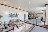 Located at 196 W Vine St in Murray, Utah, this well-appointed single-floor community space offers a generously sized, open layout designed for comfort and functionality. The living area is expansive, featuring multiple distinct seating zones furnished with a mix of modern and mid-century inspired pieces, including plush sofas, accent chairs in earthy greens and grays, and wood coffee tables. A decorative fireplace with paneled millwork serves as a central focal point, topped by a classic wall clock and flanked by warm sconce lighting. Crown molding and recessed lighting run throughout, adding a refined architectural touch. Large windows along the perimeter flood the space with natural light and provide views of the surrounding area. Thoughtfully curated artwork, including a striking equestrian painting and a vibrant landscape piece, adds visual character to the neutral-toned walls. The kitchen area is well-equipped, featuring dark espresso cabinetry with glass-front uppers, white subway tile backsplash, light countertops, stainless steel appliances including a full-size refrigerator and microwave, and warm wood-look flooring that contrasts the carpeted living zones. The overall ambiance is welcoming, polished, and versatile, suitable for gathering and everyday use.
