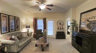 Located at 859 S Cliffs Cir in Spring Lake, North Carolina, this single-floor home offers a cozy and well-appointed living environment. The living room serves as the heart of the home, showcasing soft carpeted flooring and warm, neutral-toned walls that create a welcoming atmosphere. A ceiling fan with a built-in light fixture is centered overhead, providing both comfort and illumination. Natural light filters in through blinds and draped windows, including a glass-paneled door that connects the space to the outdoors. The room is thoughtfully arranged with plush seating, a rustic wooden coffee table, and decorative accents including an oversized wall clock and accent lamps that add character and charm. An open doorway leads to an adjacent bedroom, hinting at a well-connected and functional single-story floor plan. The overall ambiance is warm and homey, blending practical comfort with eclectic decorative touches that give the space a lived-in, personalized feel. This property presents a straightforward and comfortable layout ideal for everyday living.