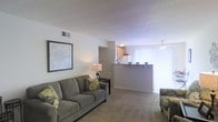 This single-floor space features an open-concept layout that seamlessly connects the living area to the kitchen. The living room is carpeted and comfortably sized, accommodating a sofa and additional seating with room to spare. A framed piece of wall art adds a decorative touch, while a small side table with a lamp provides warm ambient lighting. The textured ceiling and ceiling fan contribute to the room's casual, practical character.

A half-wall breakfast bar separates the living area from the kitchen, creating a natural division while maintaining an open feel. The kitchen features light wood cabinetry and includes a refrigerator visible from the living space. A bright window treatment allows natural light to filter into the dining or kitchen area, enhancing the overall brightness of the unit. The ceiling fan in the adjacent area ensures comfortable airflow throughout the connected spaces.

The overall ambiance is straightforward and functional, with neutral tones in the walls, carpet, and furnishings creating a cohesive, understated aesthetic. The layout is well-suited for everyday living, offering a connected flow between key living spaces.