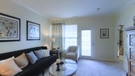 The living room serves as the focal point of this single-floor space, showcasing a thoughtfully designed interior with crown molding details that add a touch of architectural refinement. Neutral, light-toned walls create a clean backdrop that enhances the sense of openness throughout the room. A large window dressed with sheer grommet-style curtains and horizontal blinds allows generous natural light to fill the space, contributing to an airy and welcoming atmosphere. The room features a dark sofa accented with an array of decorative pillows in neutral and blue tones, complemented by a light gray accent chair nearby. A round glass-topped coffee table with a metallic frame anchors the seating arrangement, resting atop a herringbone-patterned area rug that adds visual texture to the carpeted floor. A dark wood end table with a table lamp and small decorative items provides functional detail. Two large framed botanical prints hang prominently on the wall, adding character and a cohesive decorative theme. Overall, the space presents a balanced, classic aesthetic with comfortable furnishings and well-considered design elements.