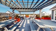 Located at 290 East Main Street in Elmsford, NY, this rooftop terrace offers a well-appointed outdoor gathering space designed for both relaxation and social engagement. The terrace is anchored by a striking pergola structure with a geometric lattice roof that casts dramatic shadow patterns across the large-format concrete tile flooring, creating a visually dynamic environment throughout the day. String lights are strung overhead, adding ambiance for evening use.

Bold orange raised planters serve as the design focal point, filled with neatly trimmed shrubs and greenery that bring life and color contrast to the space. Seating options are varied and plentiful, including classic picnic-style tables positioned beneath the pergola for shaded dining, as well as modern benches and lounge chairs arranged in open areas for more casual use. Horizontal wood-slat privacy screens and white railings define the perimeter, while still allowing panoramic views of the surrounding tree-lined neighborhood and hillside foliage.

A charming accessory structure finished in gray siding with white trim serves as an entry pavilion to the terrace, fitted with glass double doors. The overall design balances contemporary outdoor aesthetics with functional communal amenities, making this rooftop terrace a versatile and inviting space for gatherings of all kinds.