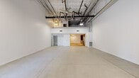 This two-floor live/work loft blends industrial character with modern comfort in a striking architectural package. The main level opens into a vast, column-free open area defined by polished concrete floors and dramatically high ceilings with fully exposed ductwork, conduit, and structural elements — an aesthetic hallmark of industrial-style design. Ceiling fans and recessed track lighting illuminate the expansive space, while large glass entry doors allow natural light to flood in and provide a visual connection to the outdoors.

Tucked behind the main open area, the kitchen is a standout feature, outfitted with high-gloss upper cabinetry in deep grey, warm wood-toned lower cabinets, a marble-veined quartz countertop and backsplash, and a full suite of stainless steel appliances including a gas range, dishwasher, and over-range microwave. A dedicated accessible restroom with grab bars is also located on the main level.

The upper level transitions to warm wood-look flooring and houses a private room with a walk-in closet offering ample built-in hanging storage. A full bathroom on this level features a glass-enclosed shower, wood-toned vanity cabinet, quartz countertop, and a large wall mirror. The two-level layout provides a natural separation between the open, flexible lower space and the more private upper quarters.