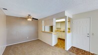 This single-floor residential space offers a practical and comfortable layout designed for everyday living. The living area features neutral-toned carpet flooring and a ceiling fan with a light fixture, providing both airflow and ambient lighting. A textured popcorn ceiling adds a classic character to the space throughout.

The kitchen is accessible via an open pass-through design, framed by a half-wall with a raised breakfast bar counter that creates a natural divide while maintaining a sense of openness between the two areas. The kitchen itself is equipped with white cabinetry, a double sink, light-toned countertops, and distinctive parquet wood flooring that adds warmth and texture underfoot. Overhead fluorescent panel lighting in the kitchen ensures a well-lit workspace.

The neutral color palette — featuring beige walls, white trim, and light-toned surfaces — creates a cohesive and inviting ambiance throughout. The open flow between the living and kitchen areas makes the space feel connected and functional. The combination of carpeted living space and parquet kitchen flooring offers a clear visual distinction between zones while maintaining a harmonious overall aesthetic.