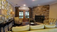 This single-floor living space combines rustic charm with traditional comfort, centered around a striking floor-to-ceiling stacked stone fireplace that serves as the room's focal point. The fireplace features a glass-front insert and warm, earthy tones that complement the overall design palette. A plush, light-colored leather sofa anchors the seating area, paired with striped upholstered accent chairs that add textural contrast and visual interest. A glass-top coffee table ties the seating arrangement together, creating a functional and stylish gathering space. Natural light enters through a set of classic wood-framed French doors, complete with divided glass panes, offering views of what appears to be an outdoor deck surrounded by lush greenery. Chocolate-brown draping curtains frame both the doors and adjacent windows, adding depth and warmth to the space. Crown molding runs along the ceiling, lending an architectural refinement to the room. Recessed lighting and a traditional table lamp provide layered illumination, enhancing the cozy, welcoming ambiance throughout. The overall aesthetic is warm and cohesive, blending natural stone, rich wood tones, and comfortable furnishings.