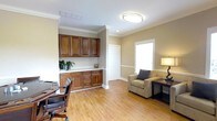 This single-floor space offers a thoughtfully designed office environment that balances functionality with a warm, professional aesthetic. The room features rich, dark wood built-in cabinetry with ample upper and lower storage, complemented by a clean white countertop workspace — ideal for organizing office essentials. Warm honey-toned hardwood flooring runs throughout, adding a welcoming feel to the environment. Crown molding along the ceiling adds a refined architectural detail, while neutral taupe walls provide a calm, neutral backdrop. The space is well-lit with a central flush-mount ceiling fixture and recessed lighting, and benefits from natural light filtering through a window dressed with blinds. A dedicated seating area features a cushioned armchair alongside a wooden desk and lamp, creating a comfortable secondary work or meeting area. A closet with paneled doors provides additional storage. The overall layout is open and well-organized, making efficient use of the available floor space. This office is well-suited for professional use, offering a clean and inviting atmosphere with practical built-in storage and versatile furniture arrangements.