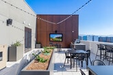 Located at 2333 Calle Del Mundo in Santa Clara, California, this rooftop terrace is a sophisticated outdoor amenity space designed for both relaxation and entertaining. The terrace is finished with elegant marble-style porcelain tile flooring throughout, lending a polished, upscale aesthetic to the open-air setting.

The space is thoughtfully organized into multiple functional zones. A fully equipped outdoor kitchen features a stainless steel built-in gas grill and stone-clad countertop, ideal for al fresco cooking and gatherings. Adjacent seating areas include a mix of high-top bar tables with stools and standard dining tables with chairs, offering flexible arrangements for casual meals or socializing.

A dedicated lounge area is anchored by a wall-mounted outdoor television set against warm wood-toned panel cladding, providing an entertainment-ready atmosphere. Raised planter beds with ornamental grasses and greenery are integrated throughout, adding natural texture to the contemporary hardscape.

String lights draped overhead create a warm ambiance during evening hours. The terrace is open to the sky, bathed in abundant natural sunlight, and enjoys sweeping panoramic views of the Santa Clara skyline, including a striking sightline to Levi's Stadium and the surrounding Silicon Valley cityscape. The overall design balances modern architecture with inviting outdoor living.