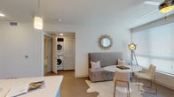 Located at 950 E Third St in the heart of Los Angeles' vibrant Arts District, this contemporary single-floor residence offers a thoughtfully designed open-concept layout finished with light wood-style flooring and a clean, neutral palette throughout. The well-appointed kitchen is a standout feature, showcasing two-tone cabinetry in white and gray, stainless steel appliances, a geometric tile backsplash, recessed lighting, and a central island with bar seating beneath elegant pendant lights — ideal for both cooking and casual dining. The kitchen flows seamlessly into the living and dining areas, which are bathed in natural light from large windows and a sliding glass door that opens to an outdoor view. A ceiling fan, cozy seating arrangements, and a marble-top dining table lend the living space a chic yet comfortable character. The bedroom is generously proportioned with plush carpeting, large windows, a ceiling fan, and access to a walk-in closet. The full bathroom features a dual-sink vanity with white quartz countertops, modern cabinetry, a large mirror, and a subway-tiled shower-tub combination. Completing the unit is a conveniently located in-unit stacked washer and dryer. This residence combines modern urban design with functional everyday living in one of LA's most dynamic neighborhoods.