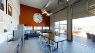 Located at 4101 South Navajo Street in Englewood, Colorado, this single-floor space showcases a compelling blend of industrial and modern design elements. The interior is defined by soaring ceilings that draw the eye upward to exposed ductwork and ventilation pipes, lending the space an authentic industrial character. A bold terracotta-red accent wall serves as a striking focal point, complemented by a large statement clock that adds both visual interest and a sense of scale to the room. The communal dining and gathering area features a long, solid-wood high-top table surrounded by contemporary metal bar stools, creating an ideal setting for collaboration or casual gatherings. Built-in bench seating lines one wall, offering additional comfortable seating options. Tile flooring throughout provides a clean, durable foundation, while modern pendant lighting contributes to the well-lit ambiance. Oversized industrial-style garage doors open to an outdoor patio area, flooding the interior with natural light and offering a seamless indoor-outdoor connection. Horizontal windows along the lower portion of the walls further enhance the brightness of the space. The overall atmosphere is energetic yet functional, making it well-suited for community gatherings, coworking, or social events.