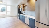This thoughtfully designed studio in Salt Lake City, UT, offers a compact yet highly functional single-floor layout that maximizes every square foot with modern finishes and smart spatial planning. The open-concept living area seamlessly integrates the kitchen, dining, and living spaces beneath bright white ceilings with recessed lighting, all unified by warm-toned hardwood flooring throughout. The kitchen is a standout feature, showcasing a sleek combination of charcoal lower cabinetry and natural wood upper cabinets and open shelving. A white tile backsplash, white quartz countertops, a black-finish pull-down faucet, and a full suite of stainless steel appliances — including a range, microwave, dishwasher, and refrigerator — make this kitchen both stylish and practical. The living area benefits from a window that invites natural light, creating an airy ambiance. A sliding barn door leads to the well-appointed full bathroom, which features light wood cabinetry with black hardware, a white quartz vanity top, a large mirror, and a glass-enclosed walk-in shower with clean white tile surround. The studio also includes a dedicated closet for storage and an in-unit laundry area, adding everyday convenience to this efficiently designed urban residence.