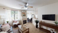 Located at 3301 E Ray Rd in Gilbert, Arizona, this single-story residence offers a thoughtfully designed layout that balances comfort and functionality. The living room serves as the heart of the home, featuring plush carpeting, a ceiling fan, and a tile-surround fireplace that anchors the space. Large windows dressed with sheer curtains flood the room with natural light and provide views of the surrounding greenery. The living area flows seamlessly into the dining space, which features a glass-top round table and corner windows that further enhance the bright, airy atmosphere. The kitchen is equipped with tall light-wood cabinetry, granite-style countertops, black appliances including a range, microwave, and refrigerator, and tile flooring. A dedicated laundry area is accessible directly from the kitchen corridor. The two bedrooms are generously sized, each featuring ceiling fans, plush carpeting, and large windows that invite natural light. The bathrooms are well-appointed with warm wood-toned vanities, granite-style countertops, and rich wood-look plank flooring. An arched hallway connects the private areas of the home, adding an elegant architectural touch. The overall design presents a warm, neutral palette throughout.