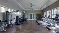This fitness center offers a thoughtfully designed workout environment suited for a wide range of exercise routines. The space features rich, dark hardwood-style flooring that runs throughout, providing both durability and a polished aesthetic. A tray ceiling with dual ceiling fans enhances airflow and adds an architectural touch to the room, while recessed lighting ensures the space remains well-lit at all times. Natural light pours in through a series of windows positioned along the upper and side walls, creating a bright and energizing atmosphere. The cardio zone is lined with a row of True treadmills, offering dedicated space for endurance training. Strength training is well-represented with a multi-station cable machine, a full dumbbell rack, weight benches, and additional resistance equipment. A wall-mounted flat-screen television provides entertainment during workouts. A modular shelving unit offers convenient storage for personal items or accessories. French doors at the rear of the space provide a welcoming entry point and allow for additional light. The overall ambiance is clean, modern, and functional, making this fitness center a practical and inviting facility for daily exercise.