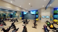 This fitness studio is thoughtfully designed to accommodate a variety of exercise routines in a bright, well-appointed environment. The space is anchored by a fleet of spin bikes arranged across a light-toned flooring surface, offering ample room for cycling sessions. Full-length mirrors line the walls, visually expanding the room and allowing users to monitor their form during workouts.

Multiple wall-mounted flat-screen televisions are positioned throughout the studio, providing entertainment and guided workout content. A dedicated wall-mounted rack displays a colorful assortment of medicine balls in varying sizes, while kettlebells are neatly organized along a rack, offering a range of resistance training options. Yoga mats and stability balls are also available, supporting stretching and functional fitness routines.

The walls feature a cool blue accent color that contrasts with the neutral tones of the remaining surfaces, lending the space a modern, energetic aesthetic. Recessed lighting throughout ensures consistent illumination across the entire room. Large glass-paned French doors connect the studio to an adjacent common area, while a door with a sidelight provides direct outdoor access, contributing to an open, airy atmosphere.