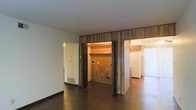This single-floor space showcases a straightforward, functional layout with notable character details throughout. The main living area features dark hardwood flooring that extends across the entire level, providing a warm and cohesive foundation. A distinctive wood-paneled accent wall serves as a central architectural element, framing a built-in storage nook or closet space with open shelving, adding both visual interest and practical utility.

The open sightlines from the main living area extend toward what appears to be a kitchen, where wooden cabinetry and a full-sized refrigerator are visible. A sliding glass or vertical blind-covered door in the kitchen area suggests access to an outdoor space, allowing for natural light to filter into the interior.

A floor vent is visible near the base of the wall in the living area, indicating a forced-air heating system. The white walls and standard ceiling height throughout the space lend a clean, neutral backdrop that contrasts with the warm wood tones. Overall, the space presents a utilitarian aesthetic with opportunities for personalization and updating.