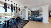 This elegantly designed single-floor communal space showcases a seamless open-concept layout that connects a spacious lounge area, dining room, and additional gathering zones. Striking herringbone-patterned hardwood flooring flows throughout, anchoring the space with warmth and visual interest.

The dining area is a standout feature, centered around a solid wood table with spindle-back chairs set atop a richly toned abstract area rug. Overhead, a dramatic cluster of elongated matte black pendant lights with gold interiors creates a bold, contemporary statement. Floor-to-ceiling windows and sliding glass doors flood the room with natural light and provide direct views and access to an outdoor pool and patio area.

The adjoining lounge is defined by a coffered ceiling with a brass geometric chandelier, a plush gray sofa, and classic accent chairs arranged around a marble-top coffee table. A dark feature wall with a stacked-wood accent adds texture and depth to the living area.

Structural white columns elegantly divide the zones without disrupting the open flow. A sleek dark island or console unit provides functional utility at the heart of the space. A glass-enclosed room visible from the main floor appears to serve as a private meeting or game room. Throughout, the neutral palette, curated artwork, and layered lighting create a refined, welcoming ambiance suited for both social gatherings and quiet relaxation.