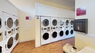This shared laundry room is a thoughtfully designed, single-floor facility built to accommodate high-volume use with efficiency and comfort in mind. The space features a generous row of stacked front-loading washers and dryers lining the walls, providing ample capacity for residents. Each machine is white, modern, and equipped with digital controls and card or coin payment interfaces, reflecting a practical, up-to-date setup.

The room is finished with warm yellow walls and durable wood-style flooring that together create a clean, welcoming atmosphere. The vaulted ceiling adds a sense of openness and height, while recessed lighting ensures the space is consistently well-lit throughout. A row of clerestory windows along the upper wall allows natural light to filter in, further brightening the interior.

A centrally positioned stainless steel folding table with lower shelving provides a functional workspace for sorting and folding laundry. Plastic bins stored beneath the table offer additional organizational utility. Informational notices are posted on the walls, and a paper towel dispenser is conveniently located near the folding area. The room also benefits from direct exterior door access, contributing to ease of use and ventilation.