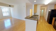This single-floor residence offers a thoughtfully designed layout that balances functionality and comfort throughout its living spaces. The open-concept living and dining area is anchored by warm hardwood flooring that reflects natural light streaming in from large windows overlooking urban surroundings, creating a bright and airy atmosphere. Clean white walls and neutral tones lend a versatile, move-in-ready aesthetic throughout.

The kitchen is a standout feature, designed in a galley style with generous cabinetry in a light honey finish and granite countertops with rich, mottled patterning. Black appliances — including a full-size refrigerator, gas range, microwave, and dishwasher — provide a striking contrast to the warm cabinetry. A peninsula with an integrated double stainless-steel sink and additional counter space connects the kitchen to the living area, ideal for casual dining or entertaining.

The bedroom offers plush carpeting, crisp white walls, and a large window that frames views of the surrounding cityscape, filling the room with natural light. The full bathroom is cleanly finished with light tile flooring, a tub and shower combination with tile surround, a toilet, and a vanity with warm wood cabinetry and a wide mirror. A hallway connects the spaces with efficient flow, completing this well-organized and comfortable residence.