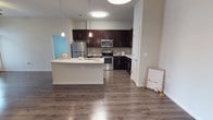 This single-floor residence offers a thoughtfully designed open-concept layout that seamlessly blends the kitchen and living areas into one cohesive, functional space. The kitchen is a standout feature, showcasing rich dark espresso cabinetry that contrasts beautifully with the light countertops and a classic mosaic tile backsplash. A full suite of stainless steel appliances — including a French door refrigerator, electric range, and over-the-range microwave — adds a sleek, modern touch. The generous peninsula with a crisp white countertop provides additional prep space and serves naturally as a casual dining or gathering area, complemented by stylish pendant lighting overhead. Wide-plank hardwood-style flooring in a warm gray tone runs throughout, grounding the space with a sense of warmth and continuity. Recessed and flush-mount ceiling lights ensure the space is well-lit throughout. Neutral wall tones keep the ambiance bright and inviting, offering a versatile backdrop for a variety of interior styles. The open flow of the layout maximizes the sense of space, making this residence feel airy and welcoming from every angle.