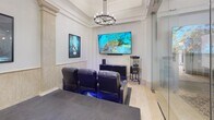 Located at 2121 S Hiawassee Rd in Orlando, Florida, this single-floor commercial space seamlessly blends upscale entertainment with professional functionality. The centerpiece of the space is a dedicated private cinema room styled after a boutique art-house theater, complete with rows of plush black leather reclining seats featuring built-in cup holders and ambient blue LED underlighting. A large wall-mounted flat-screen display serves as the main viewing screen, complemented by a sound system and a classic popcorn cart nearby. Framed movie posters adorn the walls, adding to the cinematic atmosphere. Architectural detailing throughout the theater room includes decorative stone-clad columns, wainscoting, crown molding, and elegant chandelier lighting that elevates the overall aesthetic. Adjacent to the cinema area, a stunning glass-walled conference room offers a refined and transparent environment ideal for professional meetings. This space features a large boardroom table surrounded by executive chairs, a TV display, decorative artwork, and intricate patterned tile flooring beneath elegant multi-tiered chandeliers. The open corridor connecting these areas showcases travertine-style flooring and additional chandelier fixtures, creating a cohesive and polished ambiance throughout the entire space.
