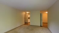 This single-floor residential space offers a straightforward and functional layout designed for comfortable everyday living. The main living area is carpeted and features neutral-toned walls that create a clean, understated atmosphere, with flush ceiling lighting providing consistent illumination throughout.

Visible from the living area, the kitchen is accessible through an open passageway and is outfitted with white cabinetry and what appears to be light-colored countertops. The kitchen benefits from its own overhead lighting and transitions into hardwood-style flooring, offering a practical and easy-to-maintain surface for a high-use area.

Adjacent to the main living space, a doorway leads to an additional room also finished with hardwood-style flooring, suggesting use as a bedroom or secondary living space. The layout connects each area in a logical flow, making navigation through the unit intuitive and efficient.

Throughout the space, the design is clean and minimal, with neutral wall colors, standard baseboards, and flush-mount lighting that emphasizes simplicity and functionality. The overall ambiance is quiet and unassuming, providing a solid foundation that can be easily personalized to suit a variety of living preferences.
