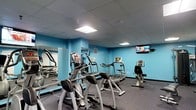 This fitness center is a dedicated single-floor space designed to support a full cardio workout routine. The room features vibrant blue walls that create an energetic atmosphere, complemented by a drop-tile ceiling with recessed fluorescent lighting that ensures the space is well-lit throughout. The flooring is covered in durable dark rubber matting, which is both practical and slip-resistant — ideal for high-traffic fitness use.

The cardio equipment lineup includes elliptical trainers, upright stationary bikes, and treadmills, all arranged in an orderly row to maximize floor space and ease of use. A wall-mounted flat-screen television is positioned at the front of the room, allowing users to stay entertained during their workouts. Large mirrors line one wall, visually expanding the space and enabling users to monitor their form while exercising. A dumbbell rack is also visible along the mirrored wall, providing access to free weights for supplemental strength training.

The overall layout is functional and thoughtfully organized, making efficient use of the available space. The combination of bold color choices, ample lighting, and a practical equipment arrangement makes this fitness center a welcoming and motivating environment for regular exercise.