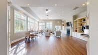Located on Cornerstone Drive in Glen Mills, PA, this generously sized club room is a well-appointed communal space designed for comfort and socialization. The single-floor layout features warm hardwood flooring throughout, complemented by soft gray walls, crisp white trim, and elegant crown molding that lend the space a refined, classic character.

Expansive floor-to-ceiling windows line multiple walls, flooding the interior with natural light and offering verdant outdoor views, including glimpses of a pool and surrounding greenery. A tray ceiling with recessed lighting adds architectural dimension and height to the main gathering area.

The room is thoughtfully divided into distinct functional zones. A lounge area is anchored by a handsome fireplace with a white mantle and flanked by built-in shelving and cabinetry, perfect for displaying décor. A wall-mounted television above the mantle adds modern convenience. Nearby, a comfortable seating arrangement with neutral upholstered sofas and armchairs rests atop a patterned area rug. A dining area with a glass-top table and upholstered chairs sits beneath large picture windows.

French doors with glass panels open to additional areas and outdoor spaces, enhancing the airy, connected feel. A service counter with dark granite and a pass-through to a brightened kitchenette area rounds out the room's amenities, making it an ideal space for gatherings and events.