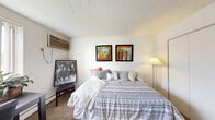 Located at 1800 Rhodes Rd in Kent, Ohio, this one-bedroom, one-bathroom unit offers a practical and efficiently arranged single-floor layout. The space is designed to maximize functionality within a compact footprint.

The spacious primary bedroom features wall-to-wall carpeting, a textured ceiling, and a wall-mounted air conditioning unit. A large sliding closet with full-panel doors provides generous storage. Natural light filters in through a window fitted with vertical blinds, creating a bright and comfortable atmosphere.

A narrow hallway connects the bedroom to the rest of the unit, leading into a versatile living and study area. This secondary space is carpeted and illuminated by a large window with vertical blinds, allowing ample natural light. The area accommodates a desk, seating, and compact appliances including a mini refrigerator and countertop microwave, making it well-suited as a combined living and kitchenette space.

The bathroom is clean and straightforward, featuring a full tub and shower combination with white tile surround, a mosaic tile floor, a toilet, and a wood-toned vanity with a white countertop. A textured ceiling and a classic gold-tone door hardware complete the classic aesthetic. The unit's overall design is simple and functional, ideal for single occupancy.