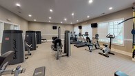 Located at 410 Starmount Park Blvd in Kannapolis, NC, this fitness center offers a comprehensive and well-equipped workout environment spread across a single floor. The space features neutral-toned walls with classic white chair-rail molding and carpeted flooring, creating a clean and comfortable atmosphere throughout.

The cardio section is well-stocked with multiple treadmills, elliptical machines, and a stationary bike, all positioned along windowed walls fitted with slatted blinds that allow natural light to filter in. Multiple flat-screen televisions are mounted on the walls, providing entertainment during workouts.

The strength training area is equally impressive, featuring a robust selection of Body-Solid selectorized machines targeting various muscle groups, a dedicated free weights station with a tiered dumbbell rack, adjustable benches, and TRX suspension training equipment. A large wall-mounted mirror spans one section of the room, allowing users to monitor their form during exercises.

Recessed lighting throughout the ceiling ensures the space is evenly and brightly illuminated at all hours. The open layout provides ample room to move between stations comfortably. The overall design balances functionality with a welcoming aesthetic, making it a practical and inviting facility for a thorough, full-body fitness routine.