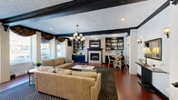 This expansive single-floor community space spans a generous footprint and is thoughtfully designed to accommodate a variety of social and functional needs. The main lounge area is a standout feature, showcasing rich hardwood flooring, bold black crown molding, and a textured ceiling that adds depth and character. A central fireplace framed by a tiled surround and flanked by built-in navy cabinetry and shelving serves as the room's focal point, while multiple seating arrangements with plush sofas invite relaxed gatherings. Elegant chandeliers and wall sconces provide warm, layered lighting throughout. Large windows allow natural light to fill the space and offer views of the surrounding exterior. A secondary lounge area provides additional seating and connects to a raised landing with decorative framed artwork. The dining area features vaulted ceilings with exposed black beams, pendant lighting, and round tables with ample seating. French doors open to an outdoor patio, further enhancing the sense of openness. A well-equipped kitchen with a bar-height counter, stainless steel appliances, mosaic tile backsplash, and granite countertops serves the dining area. A service counter area with white cabinetry and a wall-mounted TV rounds out the amenities, alongside a clean and tastefully appointed restroom.