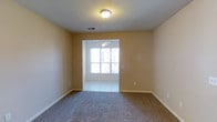 This single-floor residence offers a clean, functional layout designed for comfortable everyday living. The interior is finished with warm beige walls throughout, creating a neutral and welcoming atmosphere that pairs well with a variety of furnishing styles. The main living area features wall-to-wall carpet, providing a soft underfoot feel, while an adjoining space transitions to light-toned tile flooring, offering a practical surface ideal for higher-traffic areas or dining. A ceiling fan is installed in the adjoining room, contributing to efficient air circulation and comfort year-round. Natural light filters in through a window that faces outward, brightening the interior and lending an open, airy quality to the space. Flush-mount ceiling lighting provides additional illumination in the carpeted area. Visible HVAC vents are integrated into the ceiling, indicating a central air system. White baseboards frame the perimeter of each room, adding a crisp, finished look to the overall aesthetic. The open passage between the two areas creates a connected flow, making the layout feel cohesive and spacious despite the single-story footprint. Overall, the space presents a straightforward and well-maintained interior ready for occupancy.
