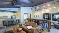 This single-floor space offers a well-designed, open-concept layout that seamlessly blends comfort and functionality. The main lounge area is anchored by a rich brown leather sofa and a wooden coffee table, creating an inviting spot for relaxed conversation or casual gatherings. Decorative accents, including colorful throw pillows and a woven bowl centerpiece, add warmth and personality to the seating area.

A long bar-height communal table with industrial-style metal stools runs along one side of the room, providing additional seating for dining, working, or socializing. The wall above is adorned with a curated series of framed artwork in cool blue tones, contributing to the space's modern aesthetic. Track lighting and strung Edison-style bulbs line the ceiling, offering both ambient and accent illumination beneath the bold, dark-painted exposed beam ceiling — a striking architectural detail that contrasts beautifully with the bright white walls.

Adjacent to the main lounge is a kitchenette area, visible through an open pass-through, equipped with a refrigerator and counter seating on bar stools. A doorway leads to an additional connected room, extending the overall footprint of the space. Light wood-style flooring throughout ties the entire layout together, creating a cohesive and contemporary communal environment.