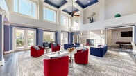 Located at 150 Coveside Lane in Chesapeake, Virginia, this generously sized single-floor community clubhouse delivers an impressive array of amenity spaces designed for both relaxation and recreation. The centerpiece is a dramatic, double-height living room adorned with a coffered ceiling detailed in navy and white, a ceiling fan, and an abundance of large windows and French doors that flood the space with natural light and provide views of the surrounding grounds. Bold red and navy upholstered seating surrounds a central fireplace, creating a vibrant yet welcoming atmosphere. A dedicated home theater room offers tiered seating with plush orange lounge chairs, a ceiling-mounted projector, deep navy wall paneling with decorative molding, wall sconces, and a built-in wet bar with quartz countertops — complete with a classic popcorn machine for an authentic cinematic experience. The well-appointed fitness center features vaulted ceilings, full-length mirrors, expansive windows overlooking greenery, and a comprehensive selection of cardio and strength-training equipment. A separate billiards and game room rounds out the entertainment offerings, showcasing a full-size pool table, shuffleboard, a mounted flat-screen TV, bold patterned carpeting, and a casual high-top seating area. Throughout, the design palette of navy, white, and warm neutrals lends a cohesive, upscale character to the entire facility.