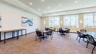Located at 2915 Bluestem Dr in West Fargo, North Dakota, this single-floor community common area is designed to serve as a flexible, multipurpose gathering space. The room is generously proportioned, with recessed lighting set into a tray ceiling that adds architectural depth and a clean, finished look throughout.

The space is divided into two functional zones. A lounge area features comfortable brown leather sofas arranged around a flat-screen television mounted on the wall, flanked by wall sconces that provide warm ambient lighting. A wooden side table and coffee table complete the seating arrangement, creating an inviting area for relaxation. A charming "Fargo" coordinates wall art adds a local touch to this section.

The opposite end of the room serves as a collaborative work and meeting area, furnished with several modular tables and mobile mesh-back chairs that can be easily rearranged. Large windows line the exterior wall, flooding the space with natural light and offering views of the surrounding building complex and outdoor grounds.

The room features neutral-toned carpet throughout, crisp white walls adorned with an abstract blue wave painting, and easy access via a secured entry door. Vending machines are visible through glass doors near the exit, adding convenience to this well-appointed community hub.