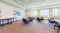 Located at 2915 Bluestem Dr in West Fargo, North Dakota, this single-floor community common area is designed to serve as a flexible, multipurpose gathering space. The room is generously proportioned, with recessed lighting set into a tray ceiling that adds architectural depth and a clean, finished look throughout.

The space is divided into two functional zones. A lounge area features comfortable brown leather sofas arranged around a flat-screen television mounted on the wall, flanked by wall sconces that provide warm ambient lighting. A wooden side table and coffee table complete the seating arrangement, creating an inviting area for relaxation. A charming "Fargo" coordinates wall art adds a local touch to this section.

The opposite end of the room serves as a collaborative work and meeting area, furnished with several modular tables and mobile mesh-back chairs that can be easily rearranged. Large windows line the exterior wall, flooding the space with natural light and offering views of the surrounding building complex and outdoor grounds.

The room features neutral-toned carpet throughout, crisp white walls adorned with an abstract blue wave painting, and easy access via a secured entry door. Vending machines are visible through glass doors near the exit, adding convenience to this well-appointed community hub.