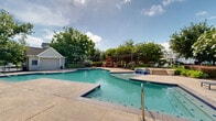 Located at 461 S Hollywood Rd in Houma, Louisiana, this single-level outdoor amenity space serves as the centerpiece of a well-appointed residential community. The highlight of the area is a generously sized, freeform swimming pool with crystal-clear turquoise water, complemented by a decorative water feature that adds a resort-like atmosphere. The expansive concrete pool deck is furnished with reclining lounge chairs, providing ample space for sunbathing and relaxation. A charming pergola structure with outdoor dining furniture offers a shaded gathering spot, ideal for socializing. Adjacent to the pool is a classic-style clubhouse building featuring a pitched roof with an arched window accent, covered porch with columns, and brick detailing — blending traditional Southern architectural character with functional design. A separate pool house or utility structure is also present near the water's edge. The surrounding landscape is lush and meticulously maintained, featuring mature flowering trees, manicured shrubs, and vibrant seasonal plantings. Decorative wrought-iron fencing and brick paver walkways frame the perimeter, creating a secure yet inviting atmosphere. The broader community grounds showcase multi-story residential buildings set against open greenspace, reinforcing the cohesive and well-planned nature of this outdoor environment.