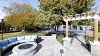 This inviting outdoor space is thoughtfully designed for both relaxation and entertainment. The centerpiece is a circular stone fire pit surrounded by a curved sectional sofa with light blue cushions, creating an ideal gathering area for evenings outdoors. The patio is finished with elegant stone pavers that extend throughout the space, providing a durable and visually appealing surface underfoot. A classic white pergola with sturdy stone pillar bases anchors one corner of the patio, adding architectural interest and a sense of structure to the open-air setting. The space is framed by mature deciduous trees with full canopies, offering natural shade and a lush, established landscape. Manicured garden beds line the perimeter, adding greenery and visual depth. A wrought iron fence borders the property, offering both security and an open sightline to the surrounding neighborhood. The combination of natural materials, thoughtful plantings, and well-chosen furnishings gives this outdoor area a warm, polished ambiance that is as functional as it is aesthetically appealing — a true extension of indoor living brought beautifully into the open air.
