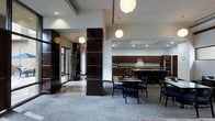 This refined single-floor space showcases a seamlessly integrated open-concept design that balances functionality with upscale aesthetics. The entrance area is distinguished by striking dark-paneled columns with horizontal accent detailing, polished marble-effect flooring, and glass-panel doors that lend a professional, corporate ambiance.

Transitioning into the main area, the space opens into a well-appointed kitchen featuring rich dark wood cabinetry that spans the full length of the back wall, a central island with a stone countertop, and stainless steel appliances. Recessed lighting and under-cabinet illumination ensure the kitchen is both practical and visually appealing.

Adjacent to the kitchen, a dining area provides comfortable seating with modern black chairs arranged around round tables, ideal for collaborative gatherings or casual meetings. The flooring transitions from the polished stone near the entrance to a neutral carpeted surface in the dining zone, subtly defining each area.

Throughout the space, contemporary globe pendant lights hang at varying heights, adding a warm and stylish focal point against the high white ceilings. Decorative greenery near the entrance softens the overall look. The cohesive use of dark wood tones, neutral walls, and modern lighting creates an environment that feels both professional and welcoming.
