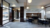 This refined single-floor space showcases a seamlessly integrated open-concept design that balances functionality with upscale aesthetics. The entrance area is distinguished by striking dark-paneled columns with horizontal accent detailing, polished marble-effect flooring, and glass-panel doors that lend a professional, corporate ambiance.

Transitioning into the main area, the space opens into a well-appointed kitchen featuring rich dark wood cabinetry that spans the full length of the back wall, a central island with a stone countertop, and stainless steel appliances. Recessed lighting and under-cabinet illumination ensure the kitchen is both practical and visually appealing.

Adjacent to the kitchen, a dining area provides comfortable seating with modern black chairs arranged around round tables, ideal for collaborative gatherings or casual meetings. The flooring transitions from the polished stone near the entrance to a neutral carpeted surface in the dining zone, subtly defining each area.

Throughout the space, contemporary globe pendant lights hang at varying heights, adding a warm and stylish focal point against the high white ceilings. Decorative greenery near the entrance softens the overall look. The cohesive use of dark wood tones, neutral walls, and modern lighting creates an environment that feels both professional and welcoming.