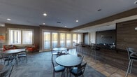 This single-floor community clubhouse is thoughtfully designed to serve as a welcoming gathering space for residents. The main gathering area is anchored by multiple round café-style tables paired with chairs, all set atop a combination of patterned carpet and tile flooring. A prominent bar-height counter lines one wall, clad in rich wood paneling and accented by stylish pendant lighting, providing an ideal spot for casual socializing or working. A large decorative clock mounted on the wood-paneled wall adds a distinctive focal point to the space.

Natural light flows generously throughout, courtesy of a wall of large windows and glass-paneled doors that look out onto an outdoor patio area, creating an open, airy ambiance. Adjacent to the main gathering area is a comfortable lounge zone, furnished with a plush blue sofa and a pair of vibrant orange accent chairs accented with patterned throw pillows, arranged around a petite side table. This seating area is further defined by interior windows that maintain a visual connection between zones. A charming vintage-style popcorn machine adds a fun, inviting touch near the lounge. Recessed lighting throughout ensures the space remains bright and inviting at any time of day. The warm, neutral color palette of taupe and gray tones lends the entire clubhouse a cohesive and modern aesthetic.