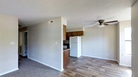 This single-floor space offers a practical and functional layout that seamlessly connects the living area to the kitchen. The living area features wood-style plank flooring that adds warmth and durability, while a ceiling fan with a light fixture provides both comfort and ambient illumination. The textured ceiling adds a classic touch throughout the space.

The kitchen is partially open to the living area, defined by a dividing half-wall that creates a sense of separation without fully enclosing the space. It features warm-toned wooden cabinetry on the upper and lower sections, dark countertops, and a full-size white refrigerator. A ventilation hood is visible above the cooking area, and the kitchen is well-integrated into the overall flow of the unit.

Neutral gray walls run throughout, providing a clean and versatile backdrop that pairs well with a variety of furnishing styles. The transition between the carpeted living space and the hard-surface kitchen floor creates a clear delineation between the two zones. A centrally located HVAC vent in the ceiling ensures efficient climate control. Overall, the space presents a straightforward, move-in-ready environment suited for everyday living.